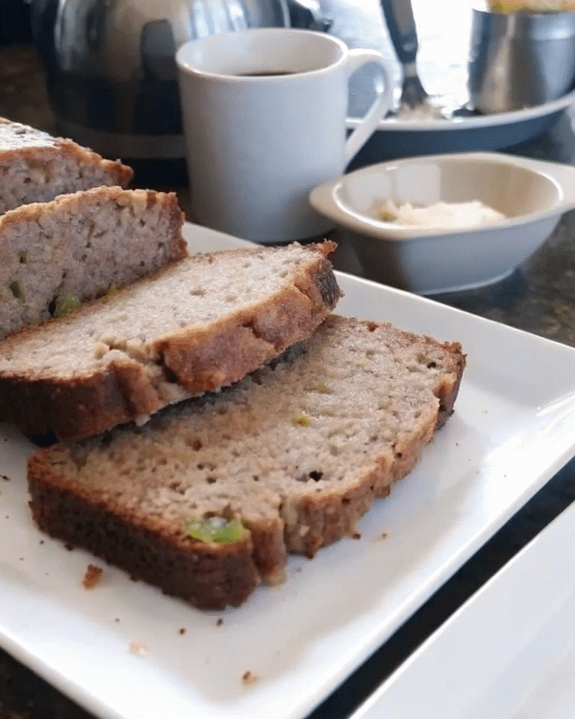 Close-up of moist zucchini bread slices stacked on a white plate. The bread has a golden-brown crust and a soft, tender interior with visible green zucchini specks.