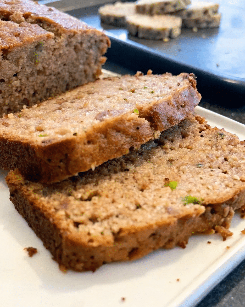 Close-up of moist zucchini bread slices stacked on a white plate. The bread has a golden-brown crust and a soft, tender interior with visible green zucchini specks.